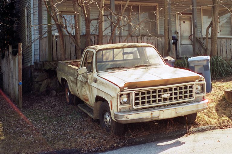 An old, rusty Chevrolet pickup truck with faded paint sits on overgrown grass in front of a weathered house, captured in a nostalgic film photo with bare trees and a blue recycling bin nearby. | Photo by Millworks Film Photography & Prints