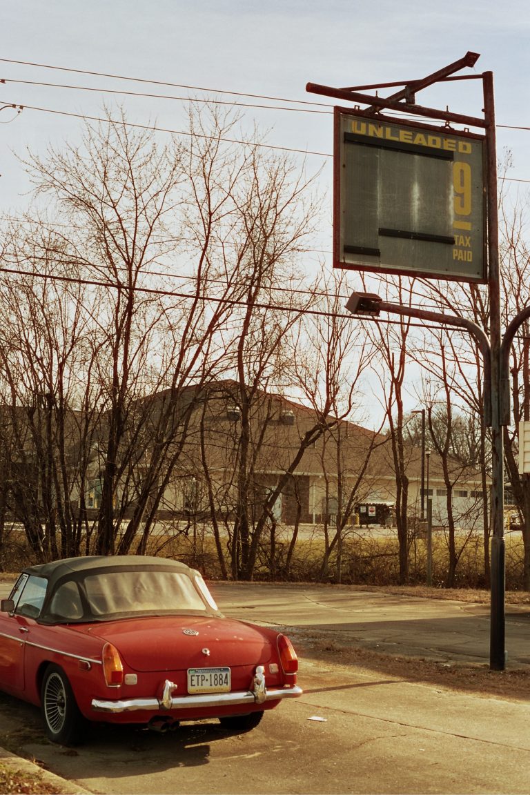 A vintage red convertible is parked near an old, empty gas station sign displaying “UNLEADED” with a faded price. Captured in classic film photography, leafless trees and buildings stand in the background under a clear sky. | Photo by Millworks Film Photography & Prints