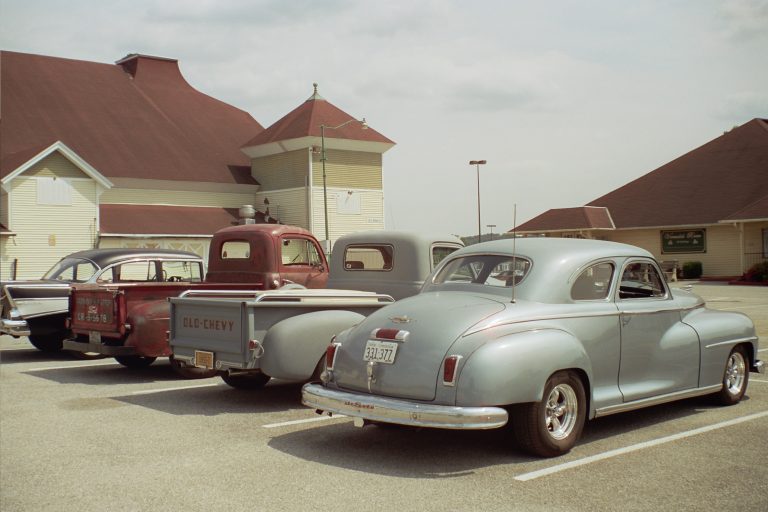 Three vintage vehicles—including two classic pickup trucks and a silver coupe—are parked side by side in a parking lot outside a building with beige siding and red roofs, captured in striking detail through professional photography on a partly cloudy day. | Photo by Millworks Film Photography & Prints