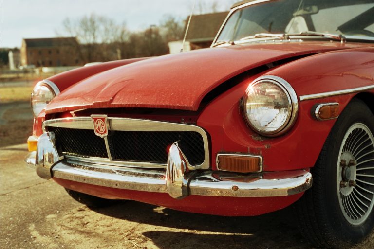 A close-up film photo of the front of a vintage red car with a damaged, slightly popped hood. The chrome bumper, round headlights, and wire-spoke wheel stand out. Professional photography captures houses and bare trees in the background. | Photo by Millworks Film Photography & Prints