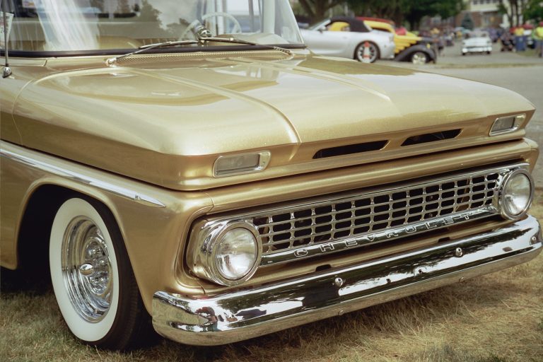 Close-up of the front end of a vintage gold Chevrolet truck with chrome details and whitewall tires, captured in classic film photography at a car show, with other classic cars visible in the background. | Photo by Millworks Film Photography & Prints