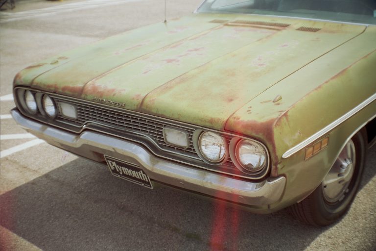 A close-up film photo of the front end of a vintage green Plymouth car with visible rust and paint fading, parked in a lot on a sunny day. | Photo by Millworks Film Photography & Prints