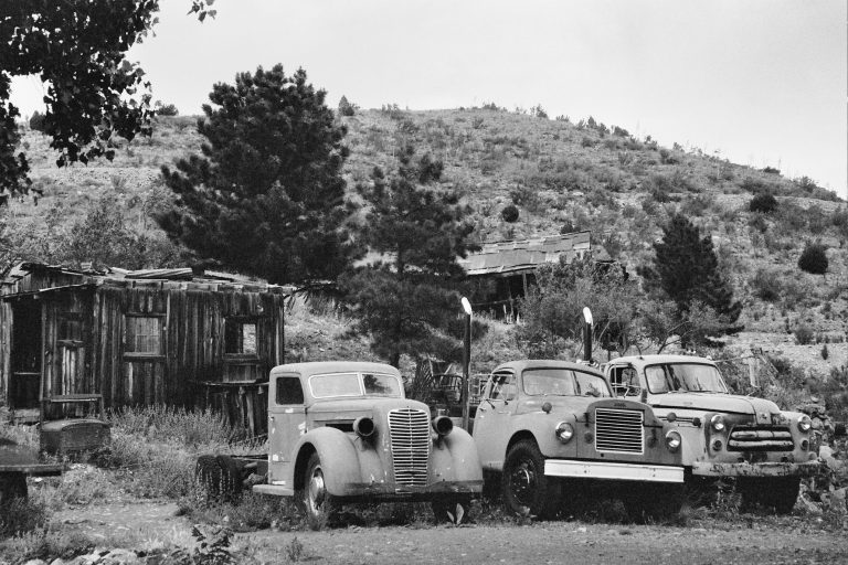 Three old, weathered vehicles sit in front of a rundown wooden shed amid overgrown grass—a film photo capturing a hilly, tree-dotted landscape beneath a cloudy sky. | Photo by Millworks Film Photography & Prints