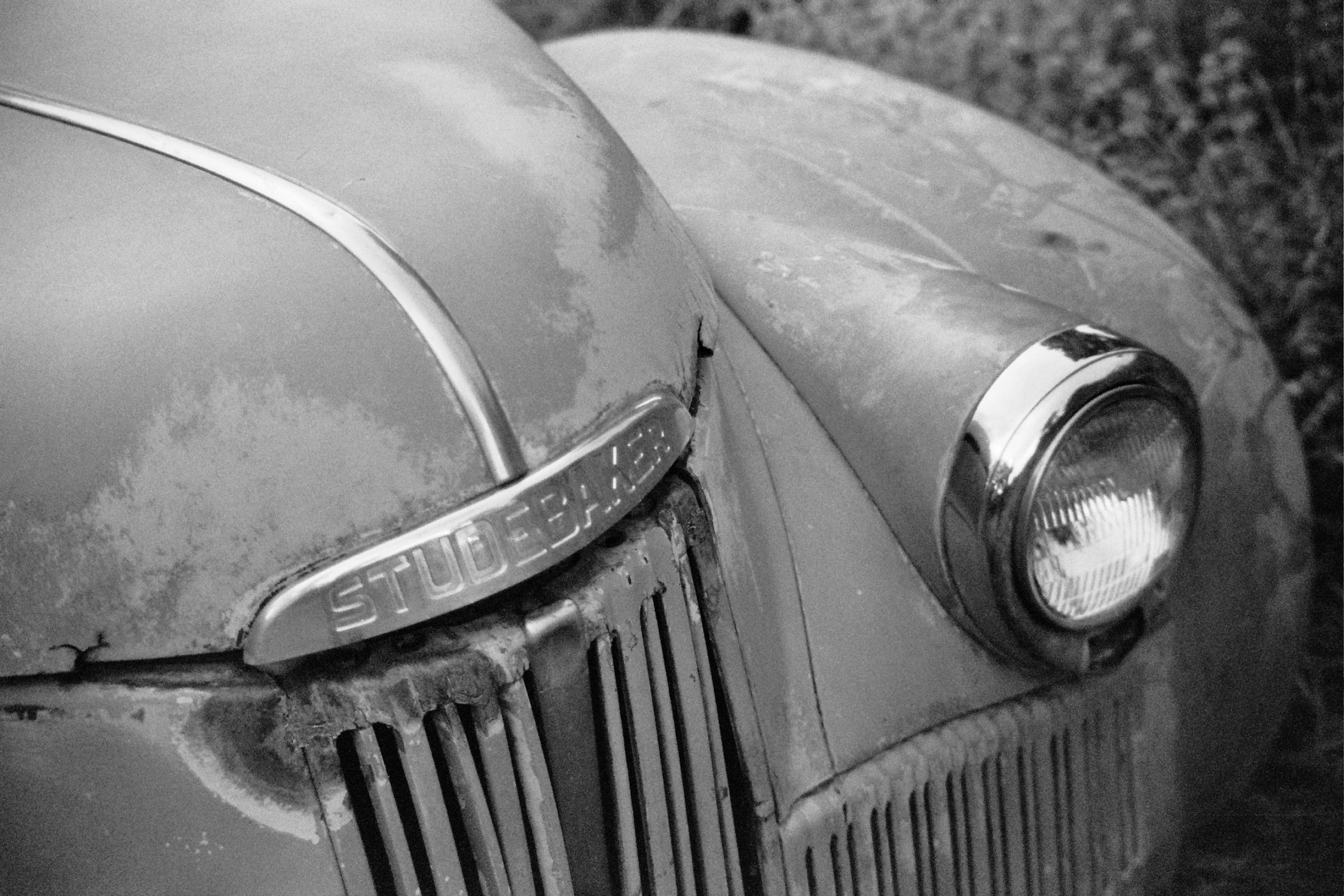 Close-up black and white film photography of a vintage Studebaker car, showing the chrome headlight, part of the hood, the STUDEBAKER badge, and a weathered, rusty grille. A true example of professional photography. | Photo by Millworks Film Photography & Prints