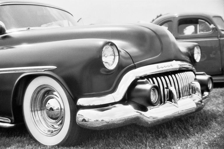A black and white film photo of a classic car, showing a close-up of the front bumper, grille, and headlight. Another vintage car is partially visible in the background, both parked on grass. | Photo by Millworks Film Photography & Prints