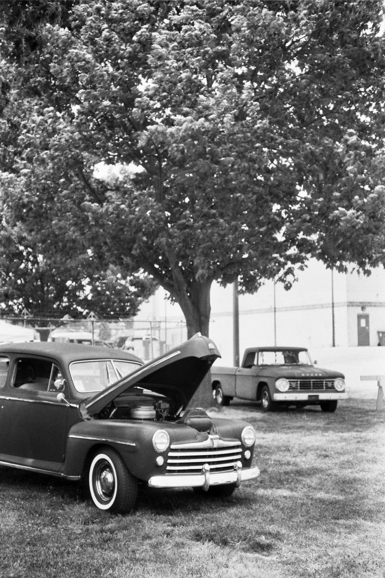 Black-and-white film photography captures a classic car with its hood open, parked on grass beneath a large tree. Another vintage car rests in the background near a building, evoking timeless style through film photo artistry. | Photo by Millworks Film Photography & Prints