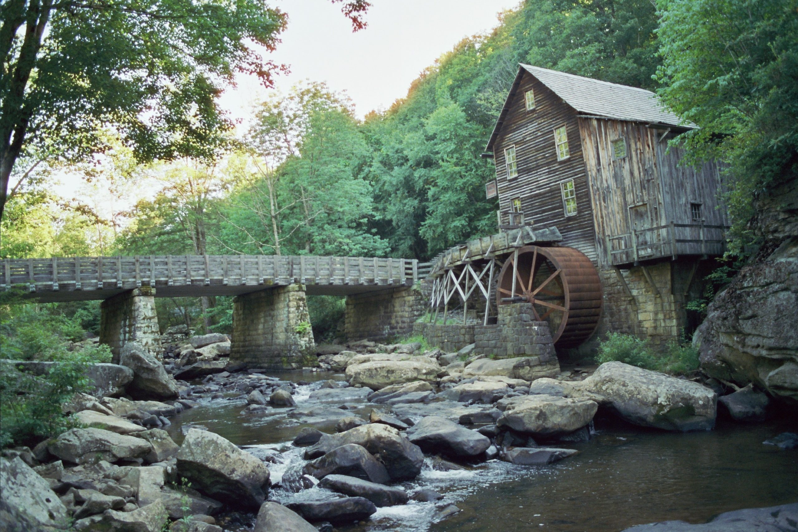 A rustic wooden watermill with a large water wheel sits beside a rocky creek, surrounded by lush green trees. Captured in the timeless style of film photography, a stone and wood footbridge crosses the creek near the watermill. | Photo by Millworks Film Photography & Prints