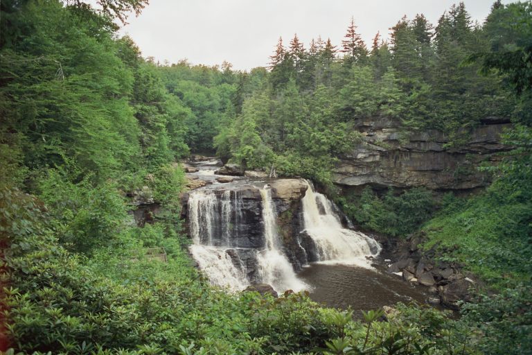 A scenic view of a waterfall cascading over rocky cliffs into a pool below, surrounded by lush green trees and dense forest under a cloudy sky, captured with the timeless charm of film photography. | Photo by Millworks Film Photography & Prints