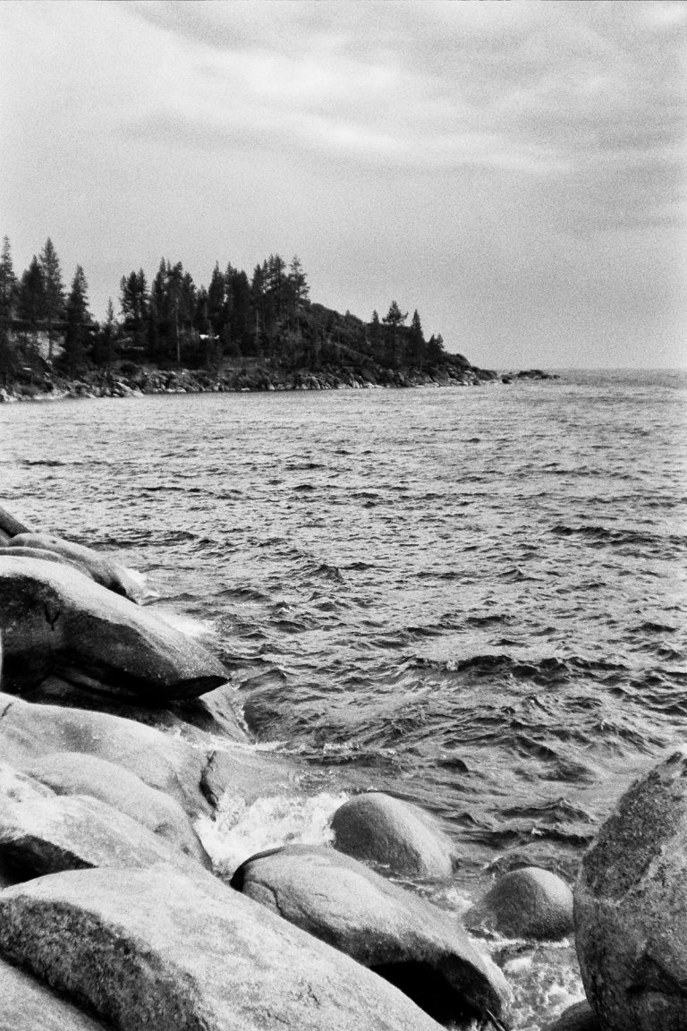 Black and white film photo of a rocky shoreline with waves hitting large stones in the foreground, a forested area in the background, and a cloudy sky overhead. | Photo by Millworks Film Photography & Prints