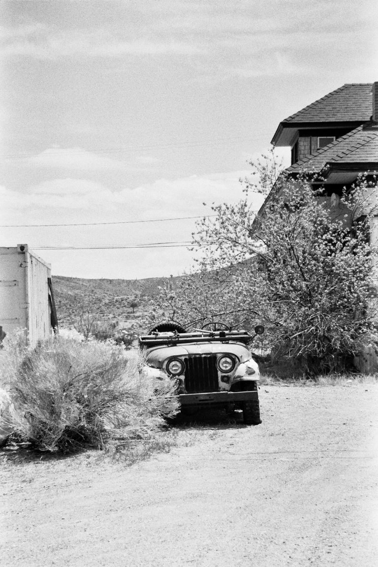 A black-and-white film photo of an old Jeep parked on a dirt road, partially obscured by a bush. In the background, there’s a building with a sloped roof and some trees, set in a rural landscape. | Photo by Millworks Film Photography & Prints