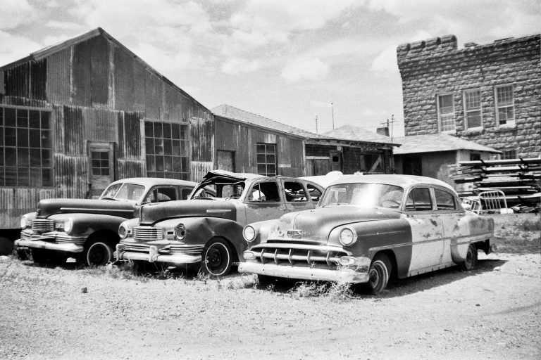 Three vintage cars with worn paint are parked on a patch of dirt in front of a rustic, corrugated metal building and a stone structure, captured in evocative film photography that highlights the nostalgic feel of this possibly abandoned location. | Photo by Millworks Film Photography & Prints