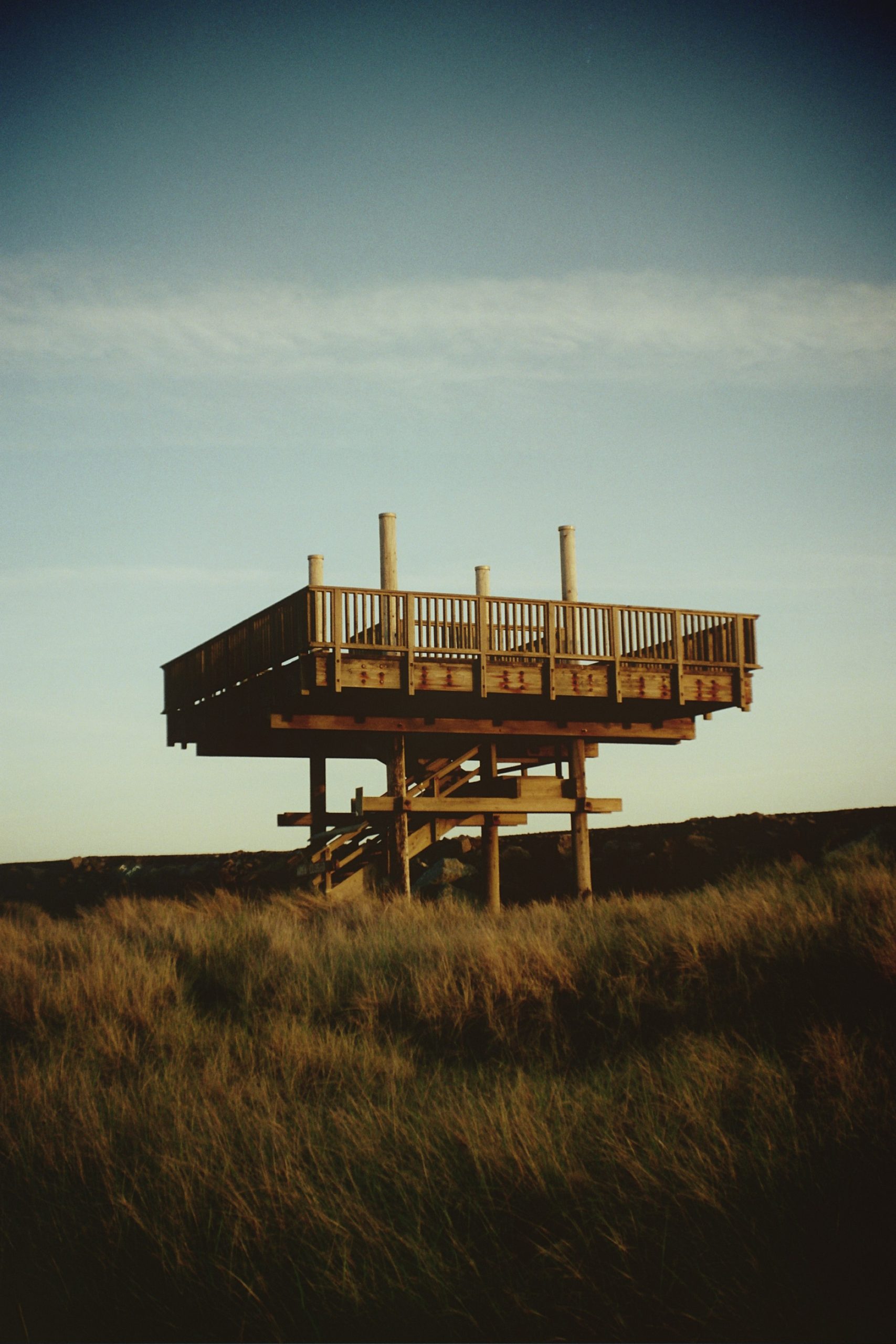 A wooden observation deck on stilts stands above tall grass in a field, with a clear blue sky in the background. Captured in stunning film photography, the scene highlights the structure’s stairs and railings wrapping around the platform. | Photo by Millworks Film Photography & Prints