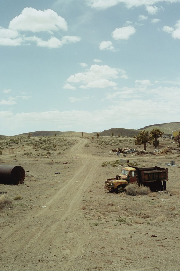 A dirt road runs through a dry, desert landscape under a partly cloudy sky. An old, rusty truck sits abandoned on the right, captured in the timeless style of film photography amid sparse vegetation and scattered debris. | Photo by Millworks Film Photography & Prints