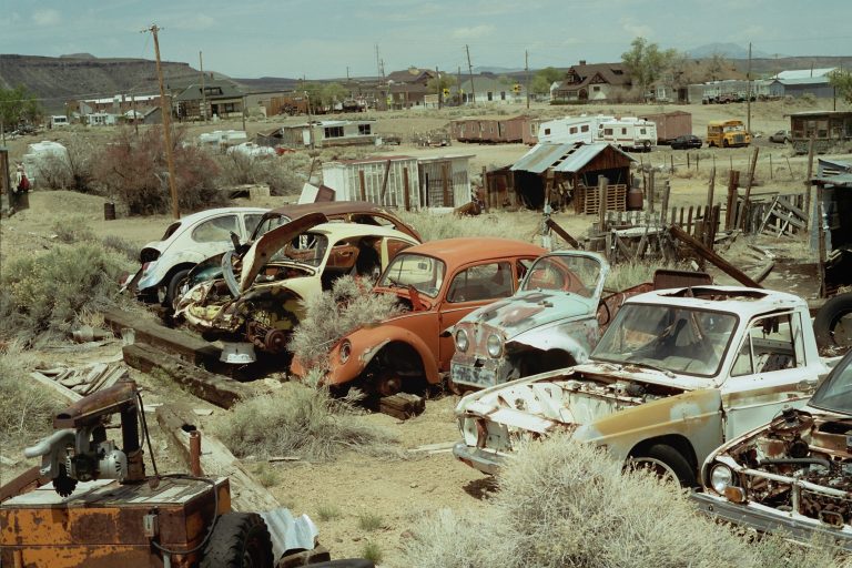 A row of rusted, abandoned cars and trucks sits among overgrown bushes in a dusty, rural junkyard with old wooden sheds and trailers in the background, captured in the timeless style of film photography. | Photo by Millworks Film Photography & Prints