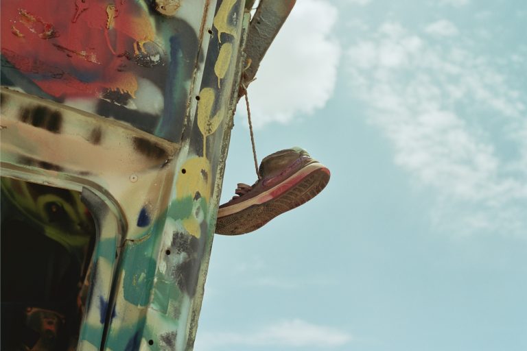 A single worn sneaker hangs by its laces from a graffiti-covered metal structure, captured in vivid detail with film photography against a bright blue sky and scattered clouds. | Photo by Millworks Film Photography & Prints