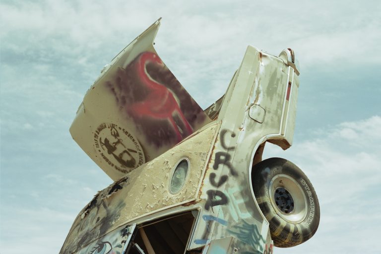 A weathered, graffitied van is partially buried upright in the ground with its back end toward the sky under a cloudy blue sky. Captured on film photography, the van features vibrant graffiti and a painted snake visible on its surface. | Photo by Millworks Film Photography & Prints