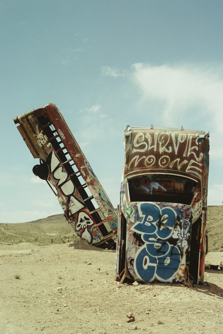Two old, rusted buses covered in colorful graffiti are partially buried upright in a barren desert landscape under a clear sky. Brown hills rise in the background, captured with the nostalgic tones of film photography. | Photo by Millworks Film Photography & Prints