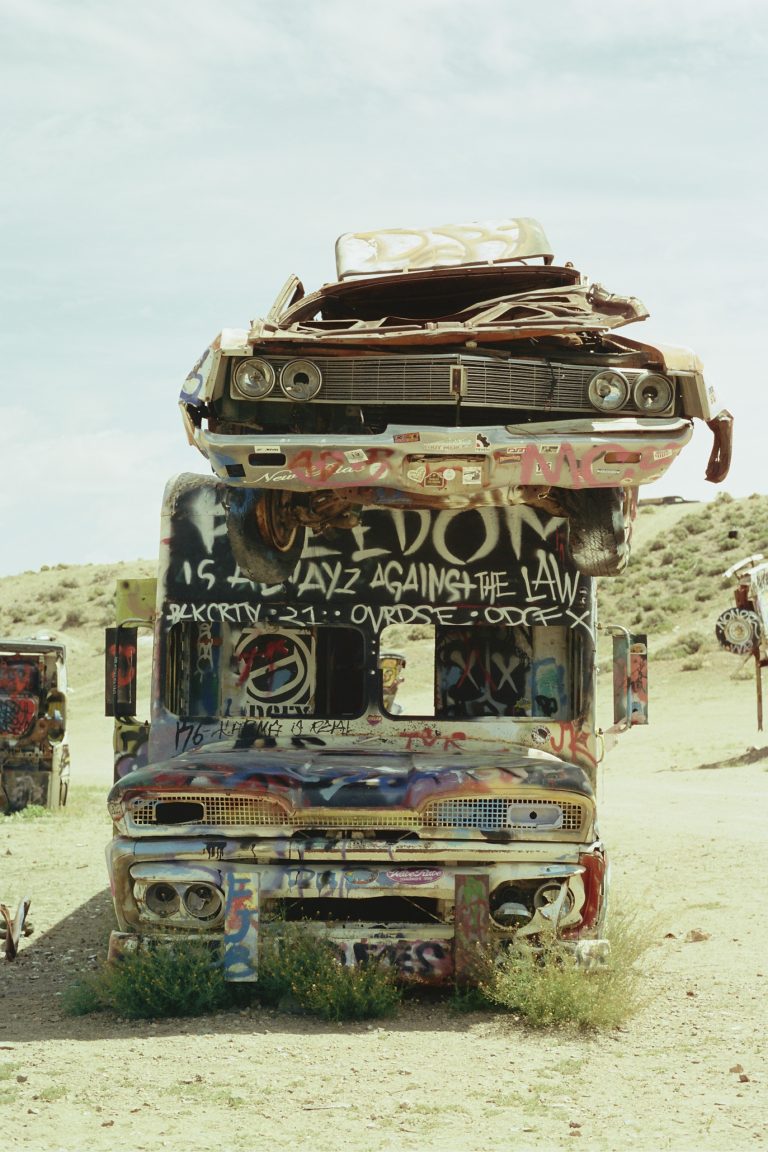 A rusted, graffiti-covered car is stacked on top of a similarly painted van in a desert landscape, captured in a striking film photo with dry grass, dirt, and clear sky in the background. | Photo by Millworks Film Photography & Prints