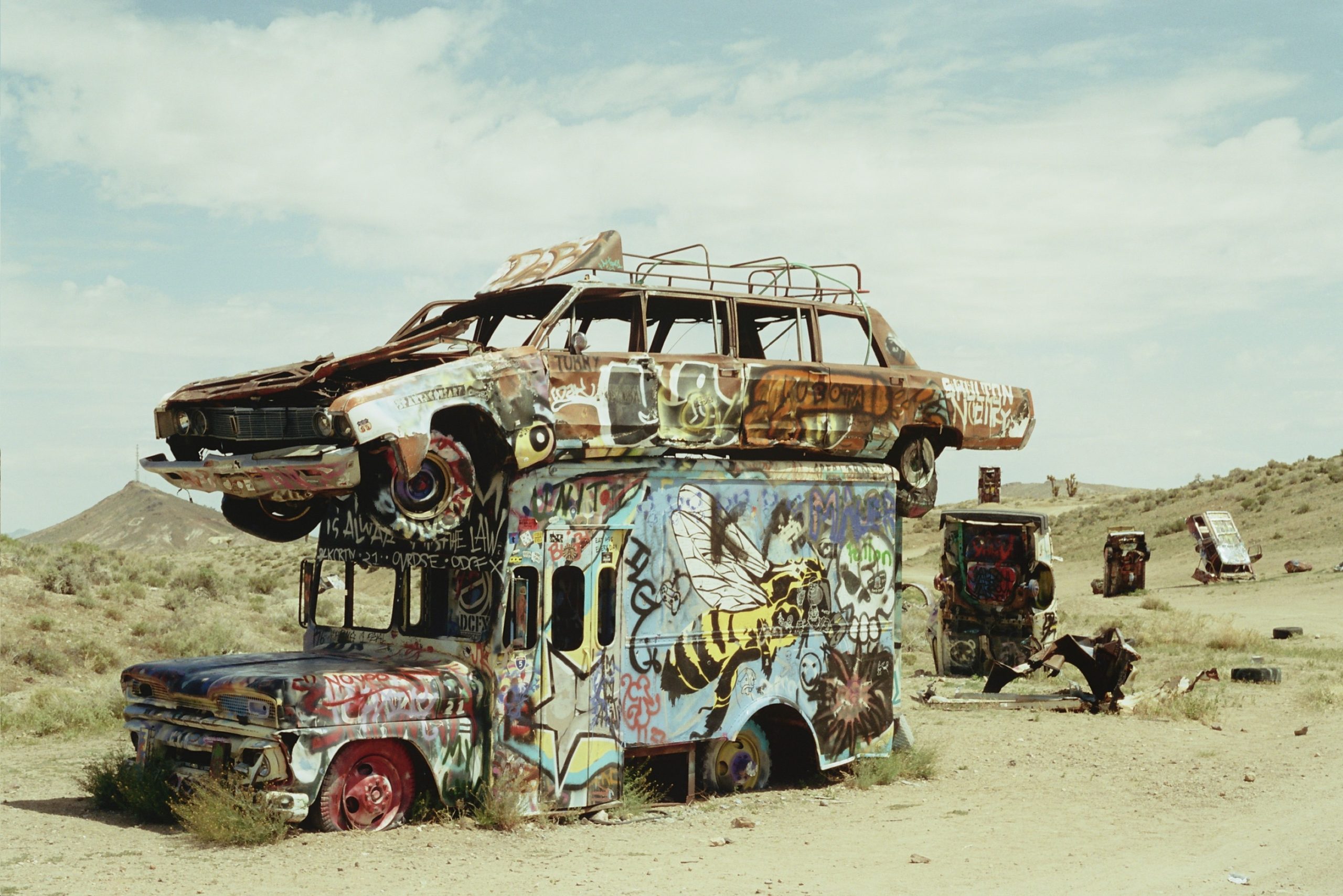 A rusted car painted with graffiti is stacked on top of a graffiti-covered bus in a desert landscape, captured in vibrant detail through film photography, with more painted, abandoned vehicles scattered under a partly cloudy sky. | Photo by Millworks Film Photography & Prints
