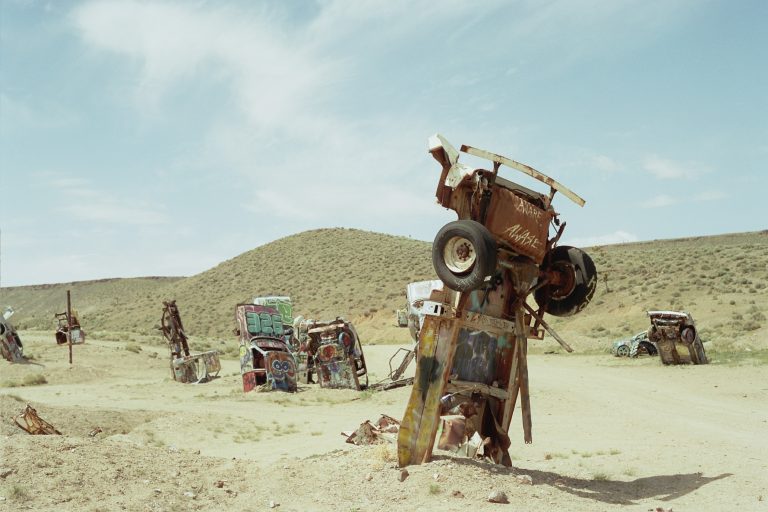 Several old, rusted cars are partially buried upright in a desert landscape, with graffiti covering some of the vehicles. Captured as a striking film photo, scrubby hills and a cloudy sky form the backdrop. | Photo by Millworks Film Photography & Prints