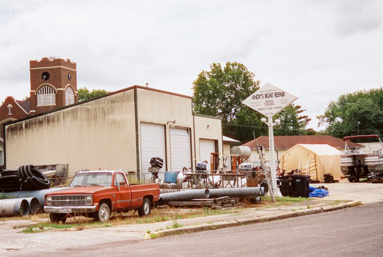 A rusty red pickup truck is parked in front of a metal building labeled “Andy’s Boat Repair.” Captured on film photography, the scene includes boats, equipment, large pipes, and a brick clock tower in the background. | Photo by Millworks Film Photography & Prints