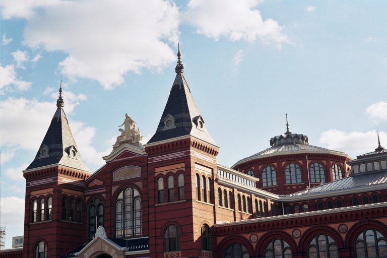 A historic red brick building with pointed slate roofs, arched windows, and ornate architectural details, captured in beautiful film photography against a blue sky with scattered clouds. | Photo by Millworks Film Photography & Prints