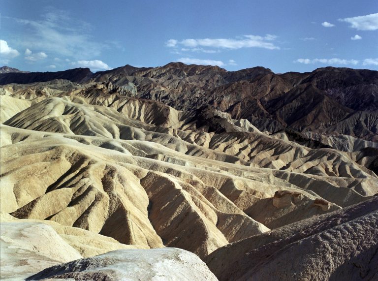 Eroded, light-colored hills and ridges with rugged, dark mountains in the background under a blue sky with scattered clouds, captured in classic film photography style. | Photo by Millworks Film Photography & Prints