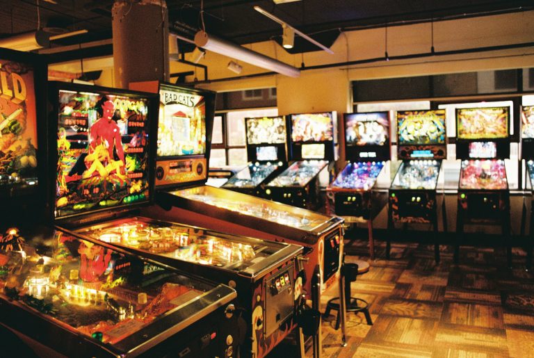 A row of colorful, illuminated pinball machines lines the floor of an arcade with patterned carpet, large windows, and vibrant themes—captured in the nostalgic style of film photography. | Photo by Millworks Film Photography & Prints