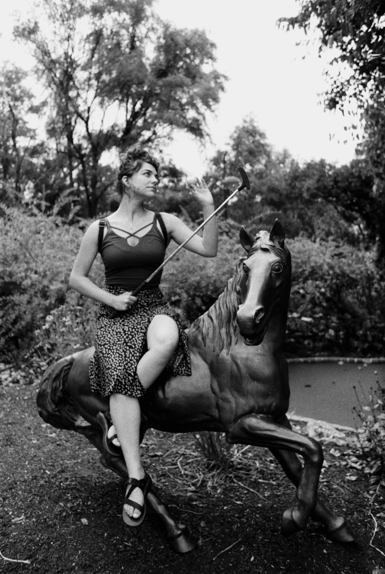 A woman in a patterned skirt and sandals sits on a statue of a horse outdoors, holding a selfie stick and looking upward. Trees and foliage surround her in the background. This black and white film photograph captures a candid moment. | Photo by Millworks Film Photography & Prints
