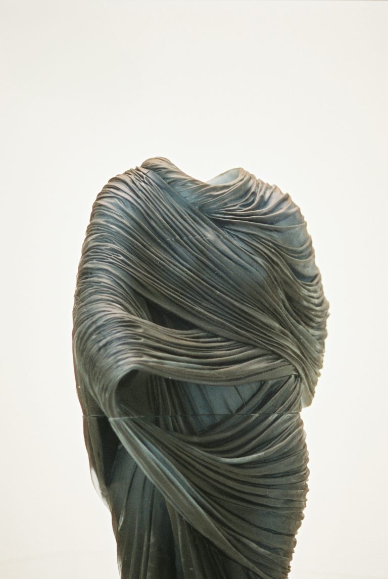 A sculpture of a human figure draped in flowing, textured fabric that obscures the face and body, with intricate folds and curves creating a sense of movement—captured in the timeless style of film photography against a plain background. | Photo by Millworks Film Photography & Prints