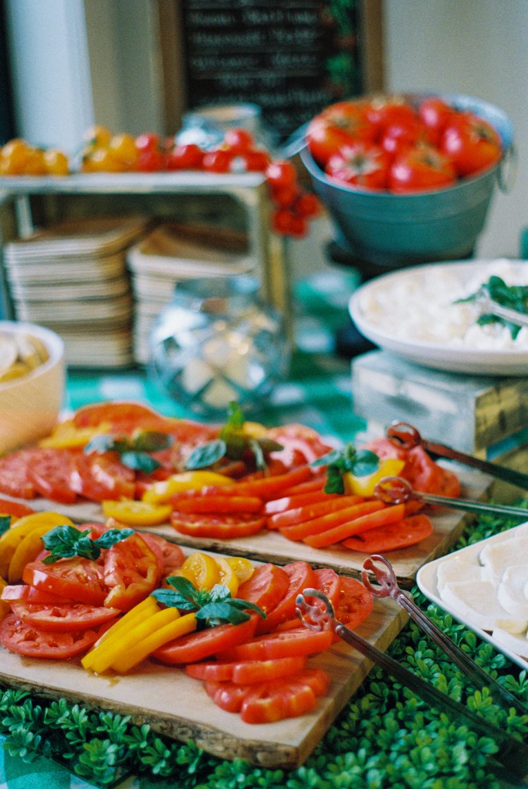 Sliced red and yellow tomatoes arranged on wooden boards, garnished with fresh basil, with bowls of cheese and buckets of whole tomatoes in the background, captured in a vibrant film photography style for a colorful, fresh food display. | Photo by Millworks Film Photography & Prints