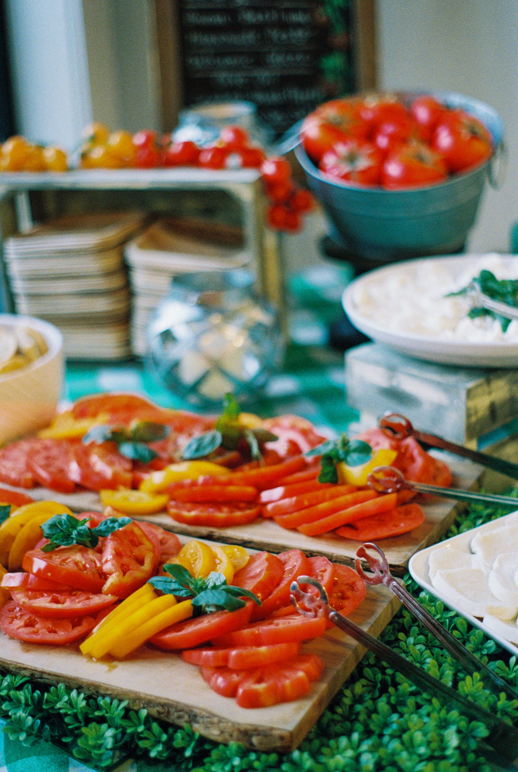 Sliced red and yellow tomatoes arranged on wooden boards, garnished with fresh basil, with bowls of cheese and buckets of whole tomatoes in the background, captured in a vibrant film photography style for a colorful, fresh food display. | Photo by Millworks Film Photography & Prints