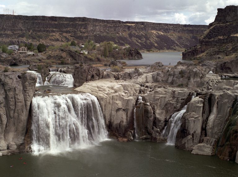 A wide waterfall cascades over rocky cliffs into a river below, surrounded by rugged terrain and sparse vegetation, with a house and hills in the background under a cloudy sky—a scene reminiscent of classic film photography. | Photo by Millworks Film Photography & Prints