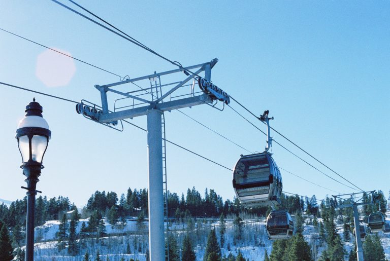 A gondola lift with cabins glides above a snowy, forested hillside under a clear blue sky in this striking example of film photography. A street lamp appears on the left side, adding character to the scene. | Photo by Millworks Film Photography & Prints