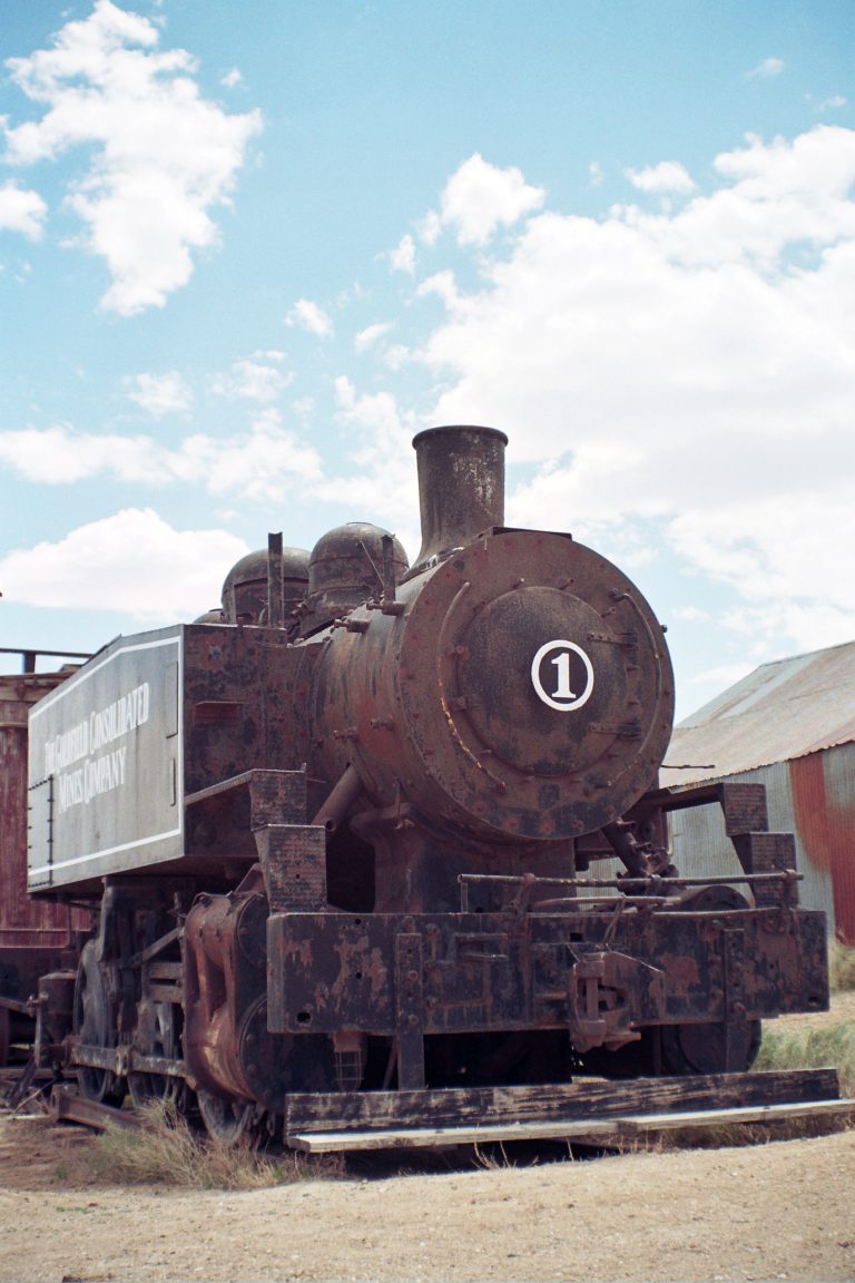 An old, rusted steam locomotive with the number 1 on its front sits on tracks in a rural area under a partly cloudy sky. Captured in the style of film photography, the faded side text reads Consolidated Mines Company. | Photo by Millworks Film Photography & Prints