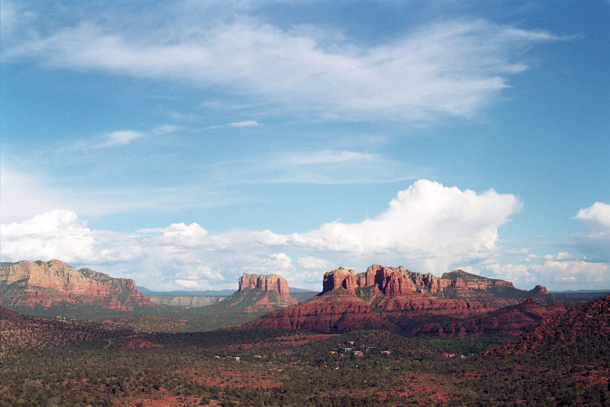 Red rock formations rise above a green desert landscape under a blue sky with scattered clouds in Sedona, Arizona, captured in striking detail through professional photography. | Photo by Millworks Film Photography & Prints