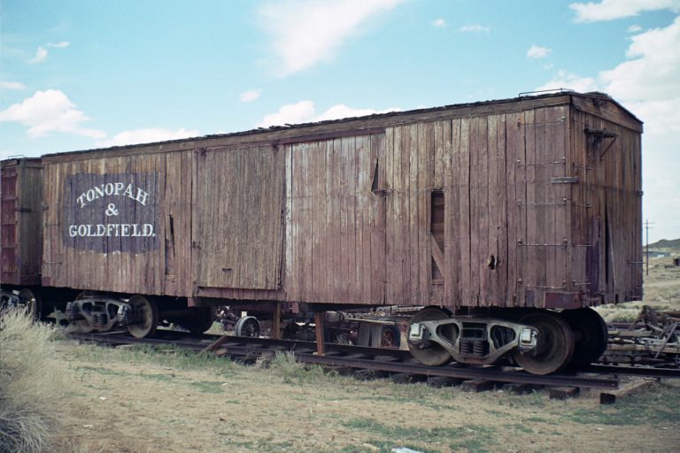 An old, weathered wooden boxcar with faded lettering reading “TONOPAH & GOLDFIELD” sits on rusty tracks in a dry, desert landscape under a partly cloudy sky, captured in timeless detail through film photography. | Photo by Millworks Film Photography & Prints