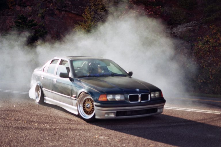 A dark green BMW sedan drifts on a road, its spinning rear tires creating a smoky cloud. Captured in film photography, the car sits low with custom wheels as rocky terrain and trees form the scenic background. | Photo by Millworks Film Photography & Prints