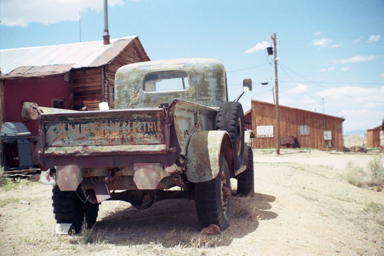An old, rusty pickup truck is parked on a dirt road beside weathered wooden buildings under a bright blue sky with scattered clouds. The scene, reminiscent of classic film photography, is framed by dry grass and sparse vegetation. | Photo by Millworks Film Photography & Prints
