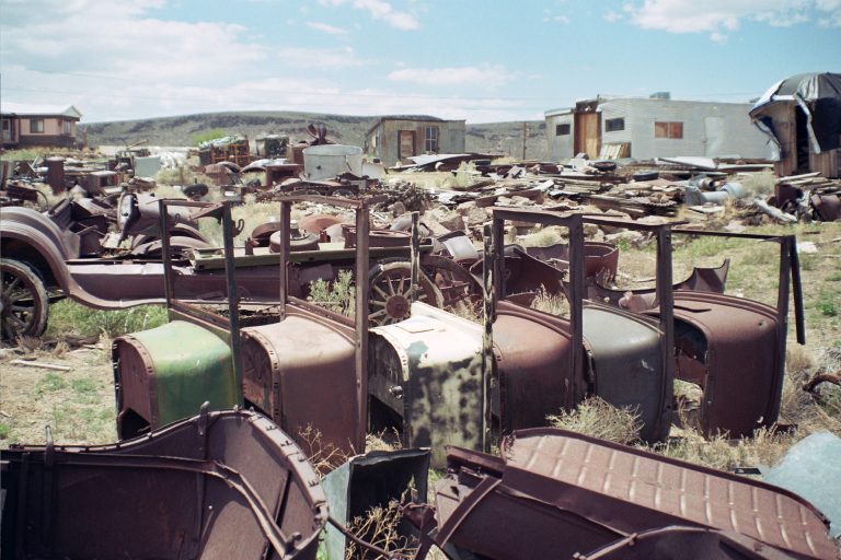 A field filled with rusted, abandoned car parts and old vehicles scattered on dry grass, captured in the nostalgic style of film photography, with weathered buildings and hills in the background under a partly cloudy sky. | Photo by Millworks Film Photography & Prints