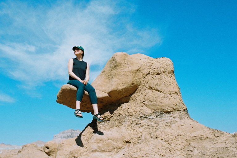 A portrait of a person wearing a black tank top, cap, and sandals sits on a large tan rock formation under a bright blue sky with wispy clouds. Distant hills are visible in the background. | Photo by Millworks Film Photography & Prints