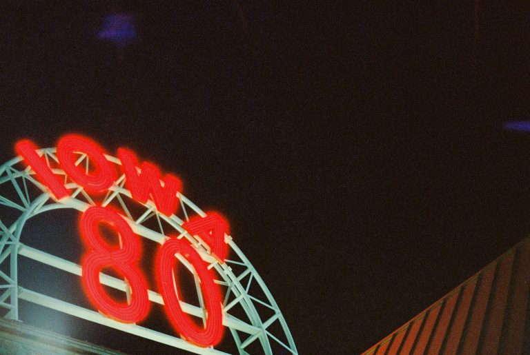 A glowing red neon sign reading Iowa 80 arches over a white metal frame at night, with a dark sky and part of a striped roof visible in the background—captured in striking detail through film photography. | Photo by Millworks Film Photography & Prints