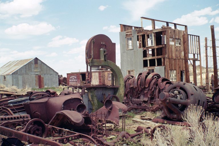 Rusty, abandoned machinery and car parts lie scattered in a field with overgrown grass beside old, weathered wooden buildings and a metal shed—captured in the timeless grain of film photography under a partly cloudy sky. | Photo by Millworks Film Photography & Prints