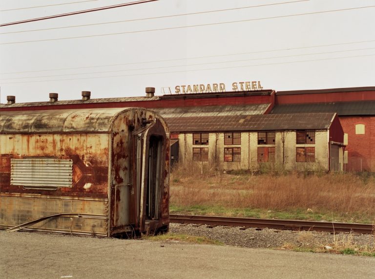 An old, rusted train car sits on overgrown tracks in front of a weathered industrial building with a faded STANDARD STEEL sign. Power lines cross the pale sky above, evoking the nostalgic mood of classic film photography. | Photo by Millworks Film Photography & Prints