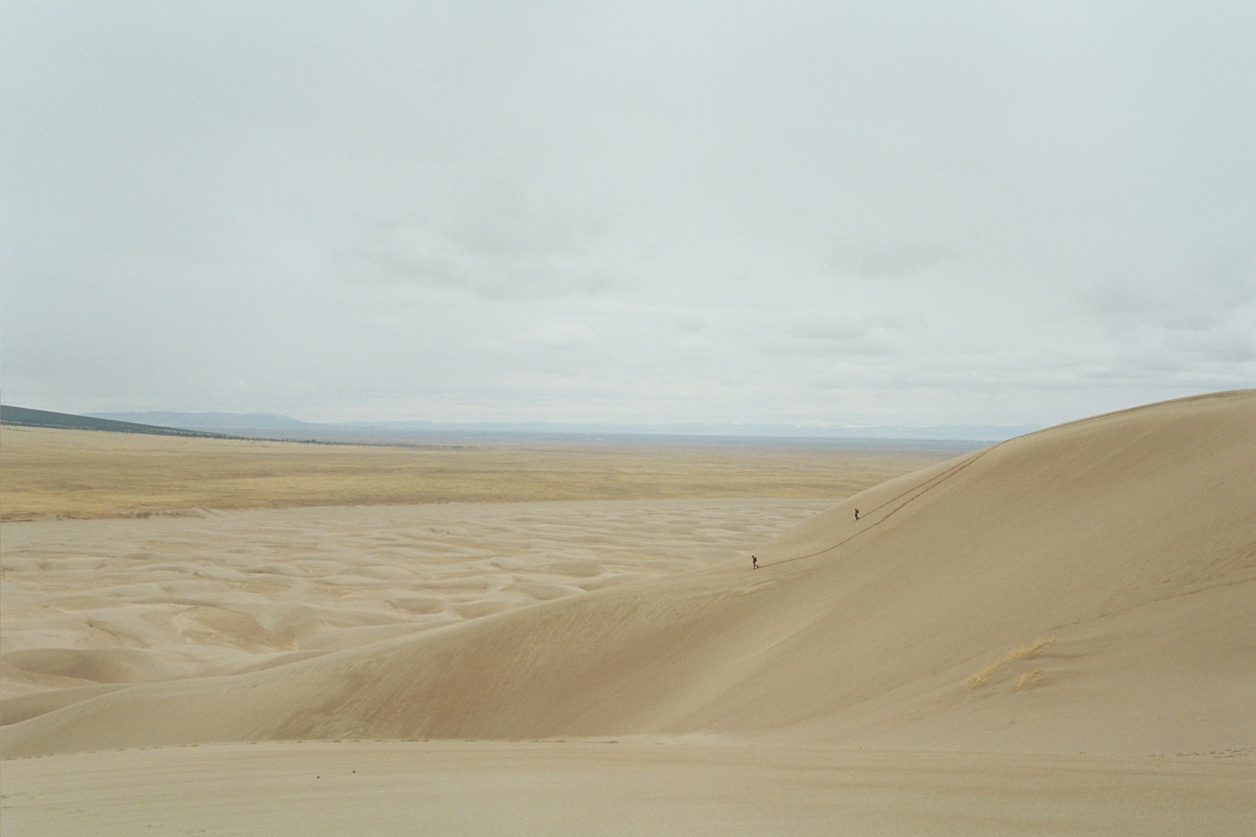 Two people walk up a large sand dune in a vast, open desert landscape under a cloudy sky, with flat plains stretching into the distance—an evocative scene captured through professional photography. | Photo by Millworks Film Photography & Prints