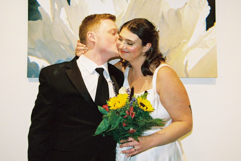 A newlywed couple poses together; the groom in a black suit kisses the bride’s cheek as she smiles, holding a bouquet of sunflowers. The warm tones of film photography highlight a large floral painting in the background. | Photo by Millworks Film Photography & Prints