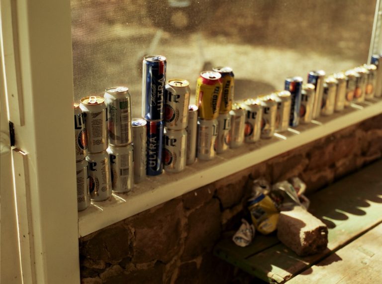 A row of empty beer cans, mostly Miller Lite and Michelob Ultra, is lined up on a windowsill inside a sunlit room with a brick wall and green wooden bench below, captured in the soft tones of film photography. | Photo by Millworks Film Photography & Prints