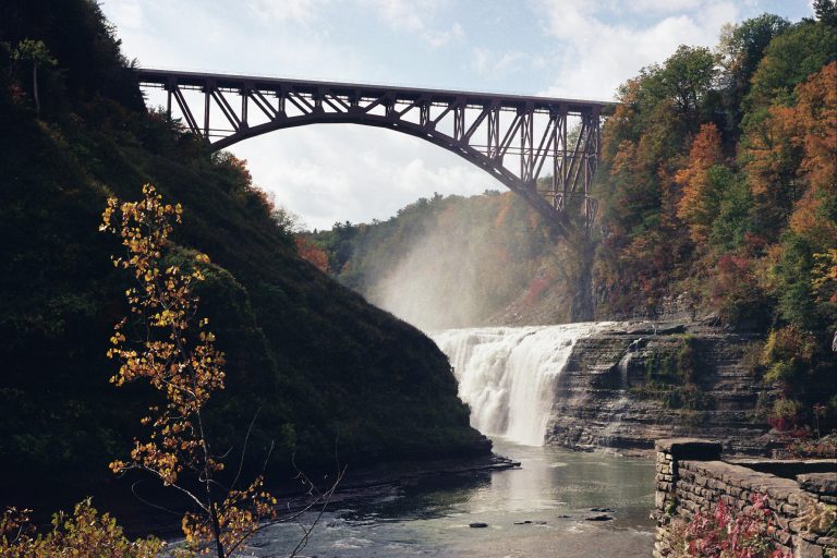 A tall steel bridge spans a deep gorge above a wide waterfall, surrounded by trees with autumn foliage. Mist rises from the waterfall on a partly cloudy day, captured in the timeless style of film photography. | Photo by Millworks Film Photography & Prints