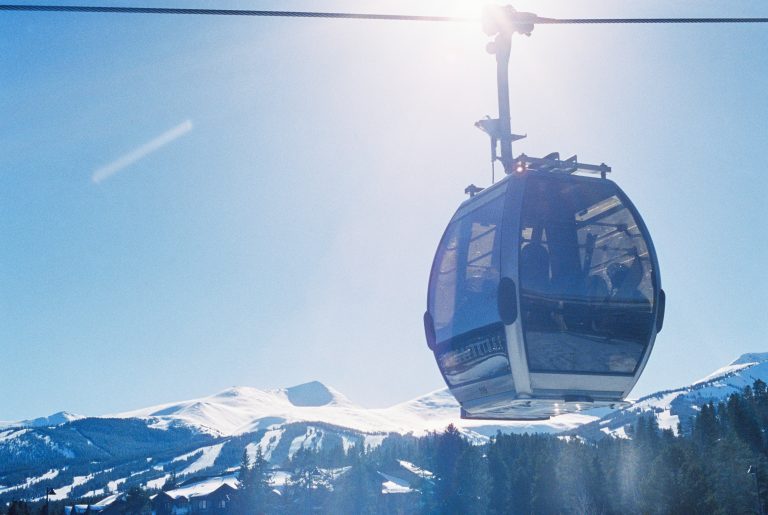 A gondola lift travels on a cable above snowy mountain peaks under a clear blue sky, with sunlight shining brightly behind the cabin, capturing the scene like a stunning film photo. | Photo by Millworks Film Photography & Prints