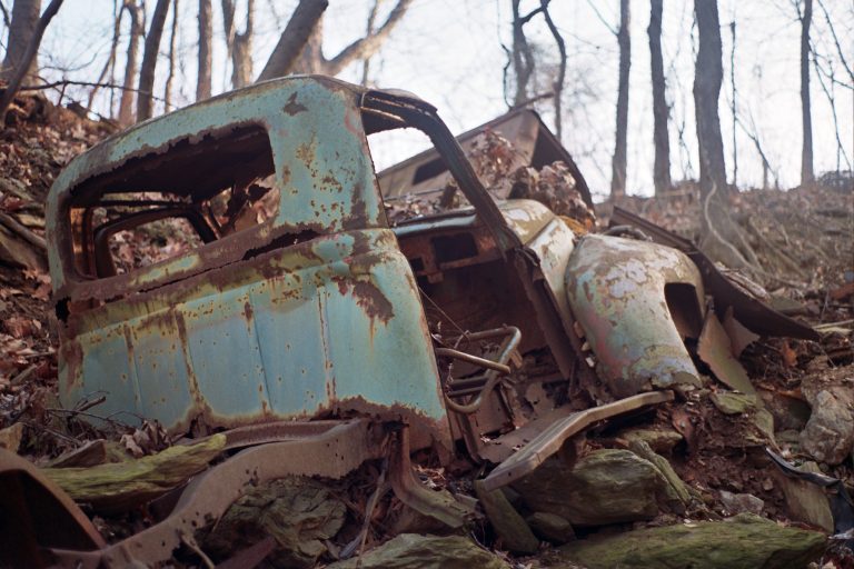 A rusted, abandoned truck sits among rocks and fallen leaves in a wooded area, with leafless trees in the background—captured through the nostalgic lens of film photography. | Photo by Millworks Film Photography & Prints
